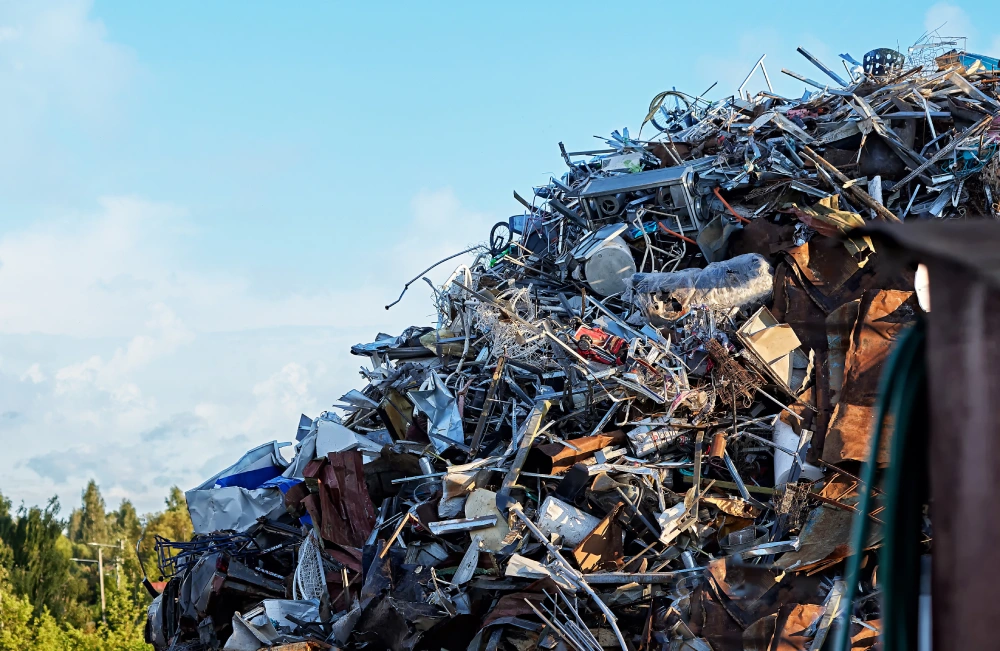 A massive mountain of mixed scrap metal debris, including wire, piping, and industrial components, towers against a bright blue sky at a recycling center.